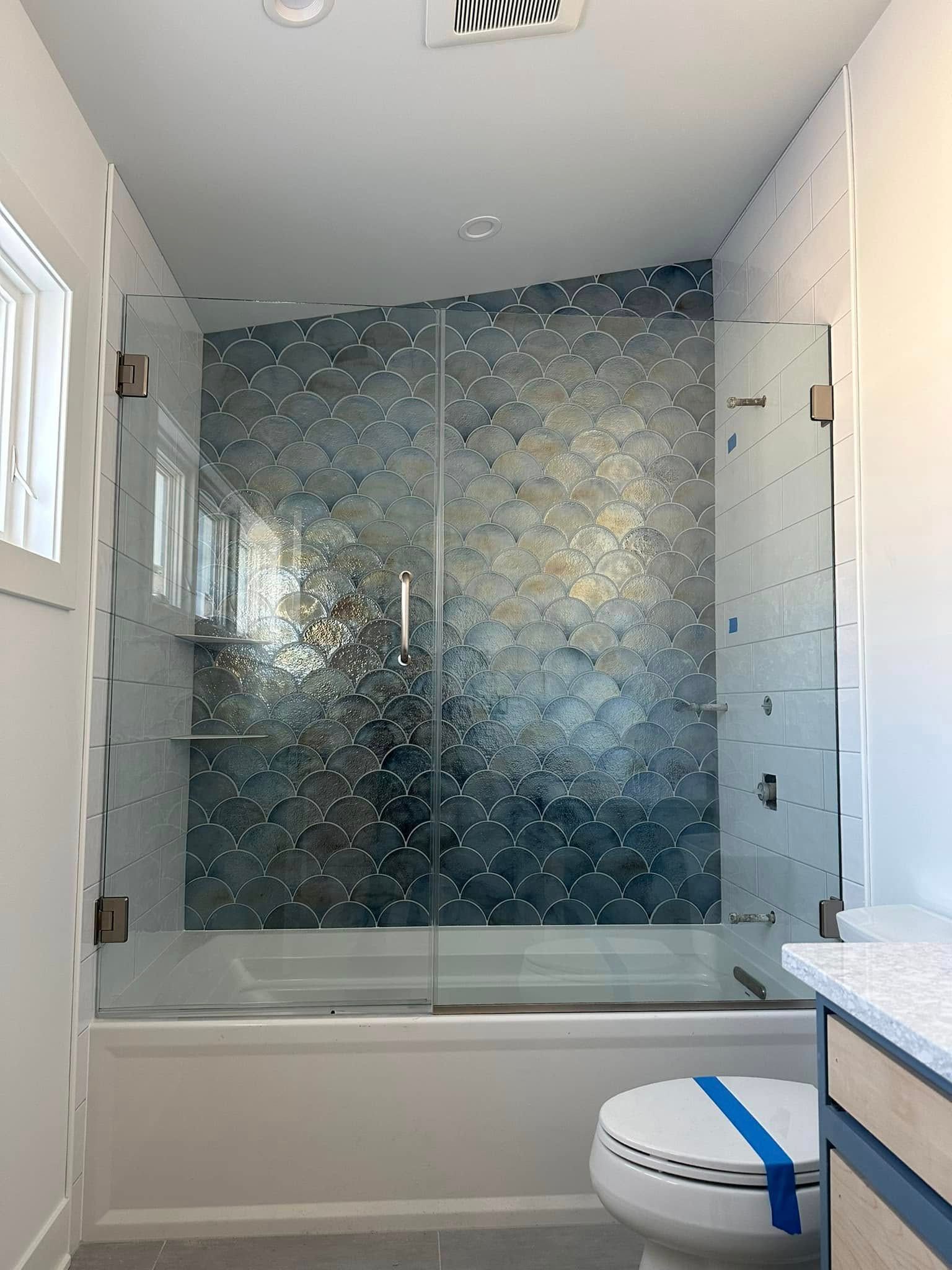A modern bathroom with a white bathtub, blue scalloped wall tiles, and a frameless glass door next to a toilet.