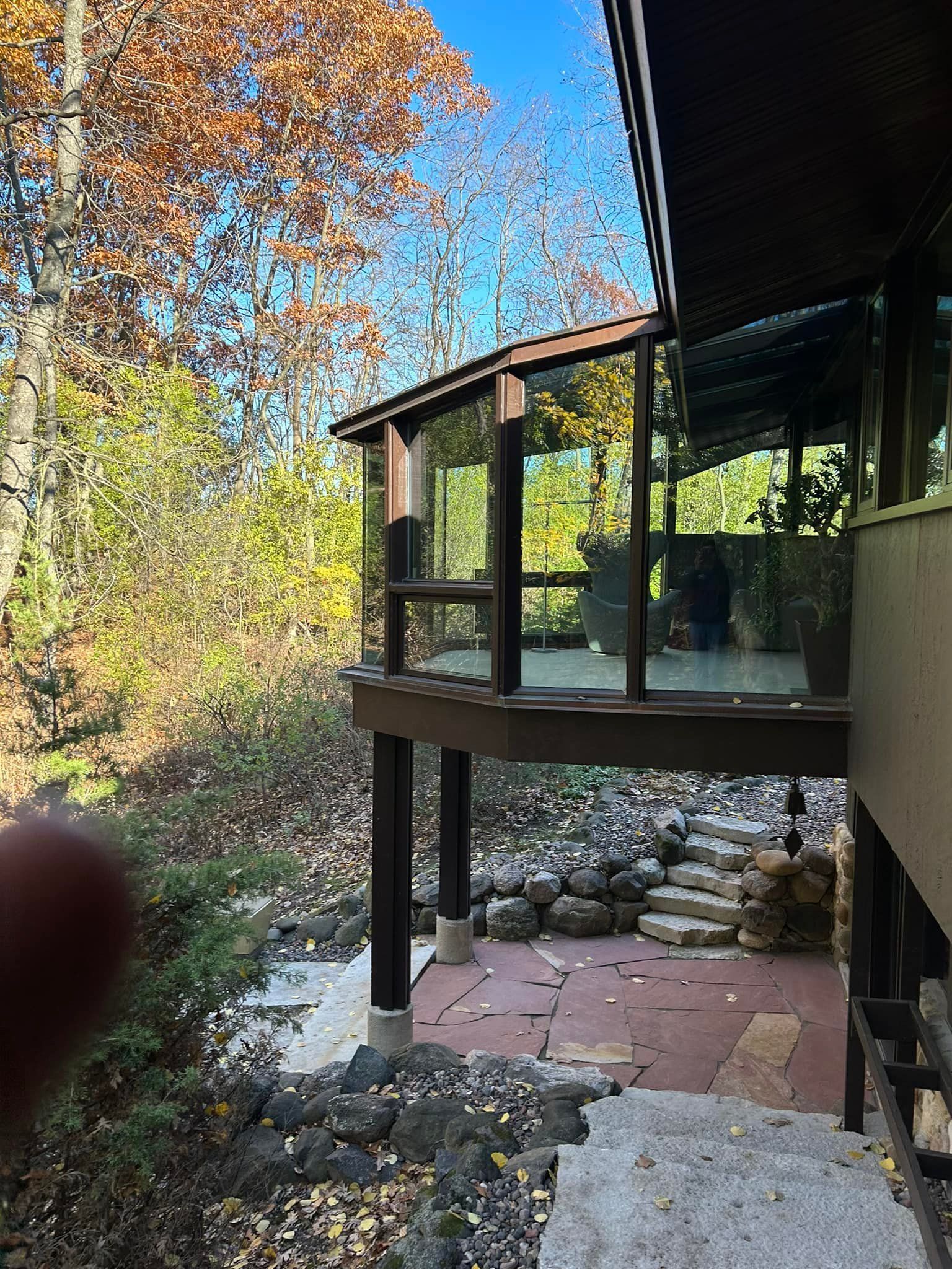 An elevated deck with glass railings overlooks a patio, stone landscaping, and trees during an autumn day.