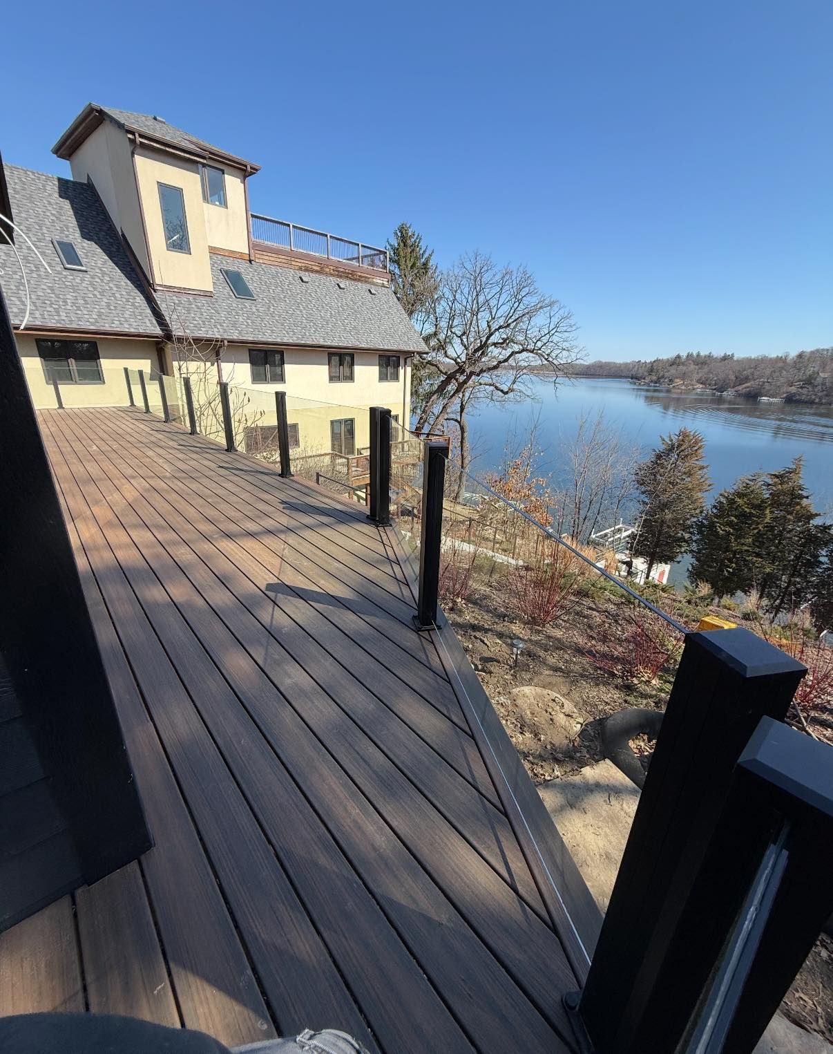 A wooden deck overlooks a large body of water and a multi-story, light-colored house under a clear blue sky.