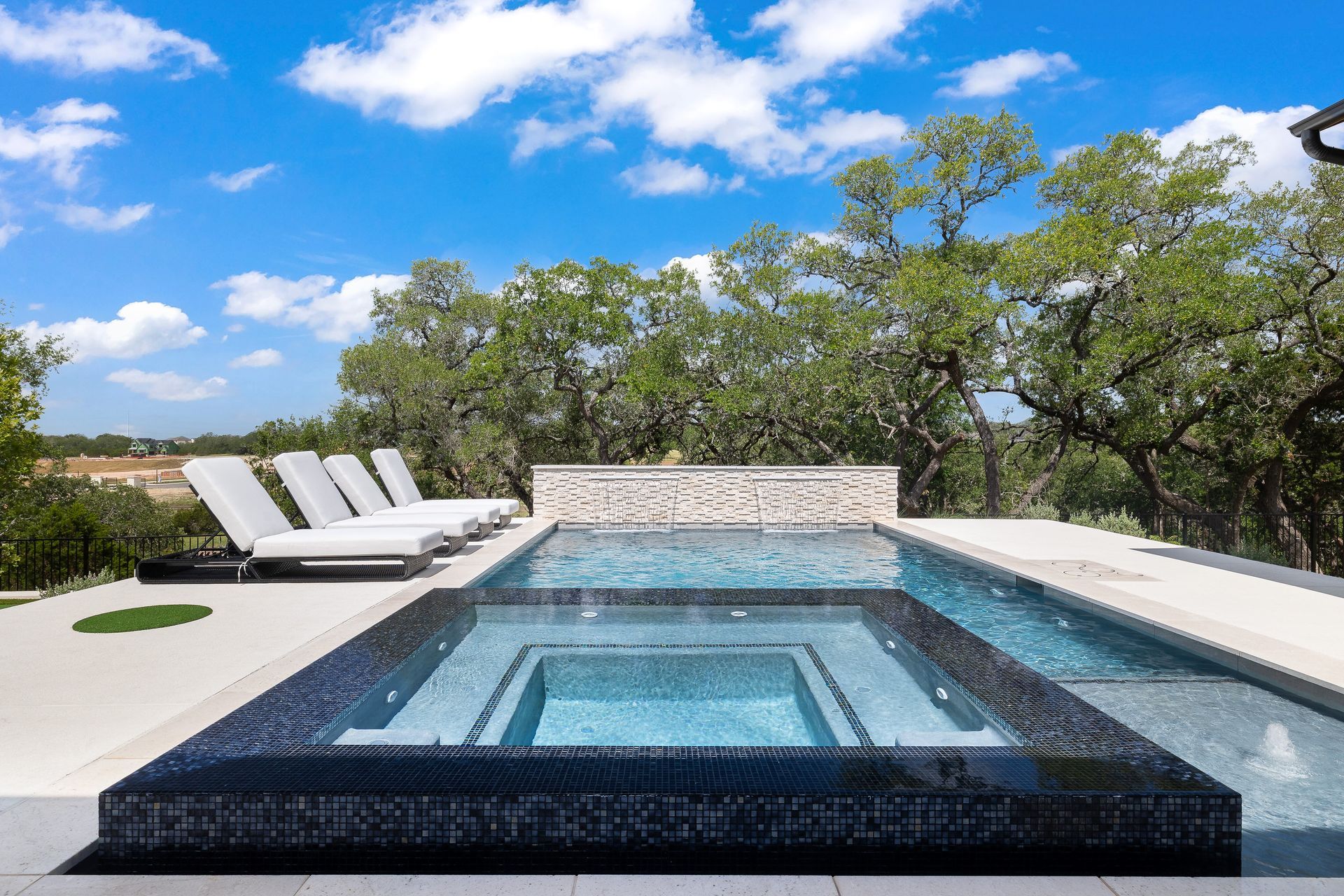 Luxurious pool with a hot tub, lounge chairs, and a scenic background of trees and sky.