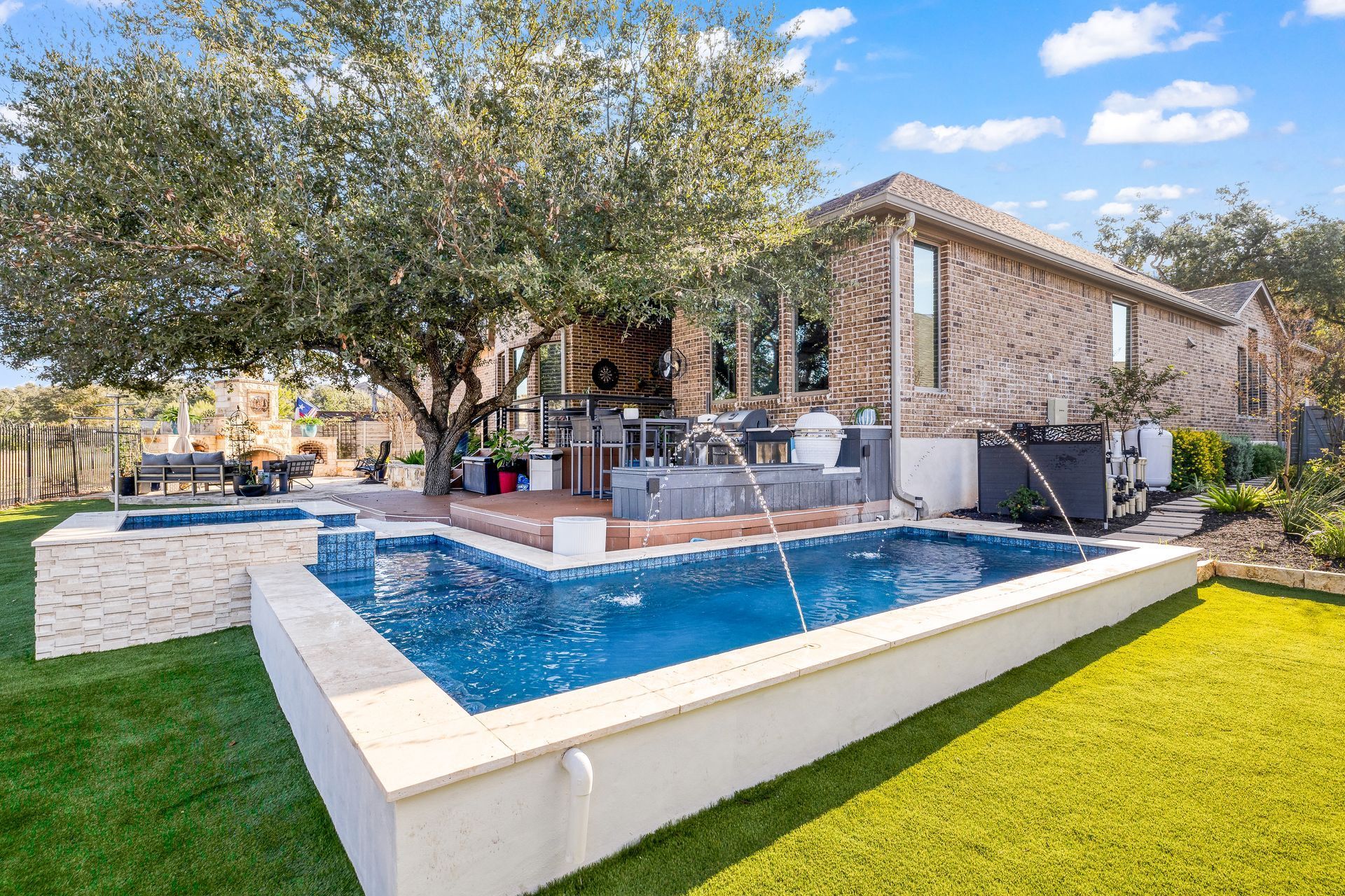 Backyard with pool, spa, patio, and brick house, surrounded by green grass and trees.