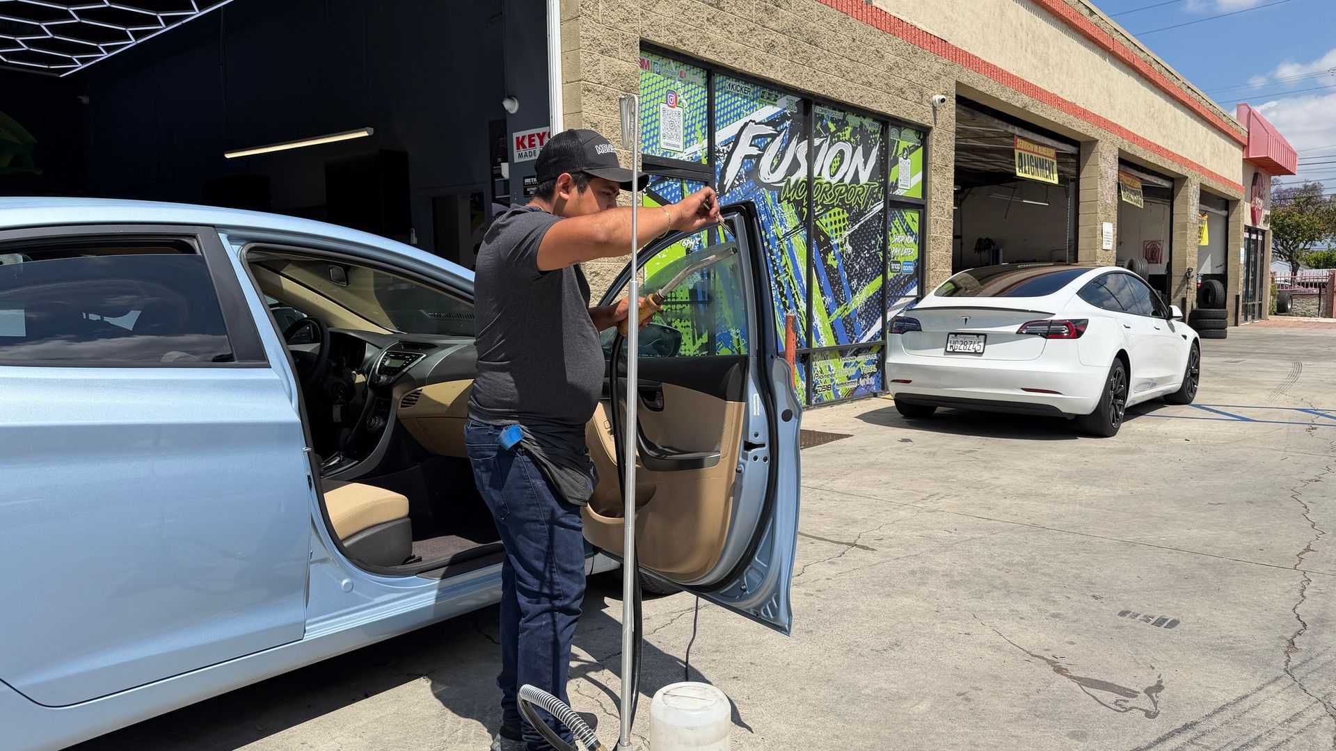Person unloading a silver car beside a storefront, holding a metal rack and blue panel on the sidewalk.