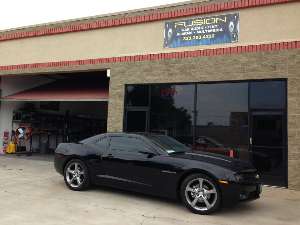 A black Chevrolet Camaro parked in front of a Fusion car audio and window tinting shop.