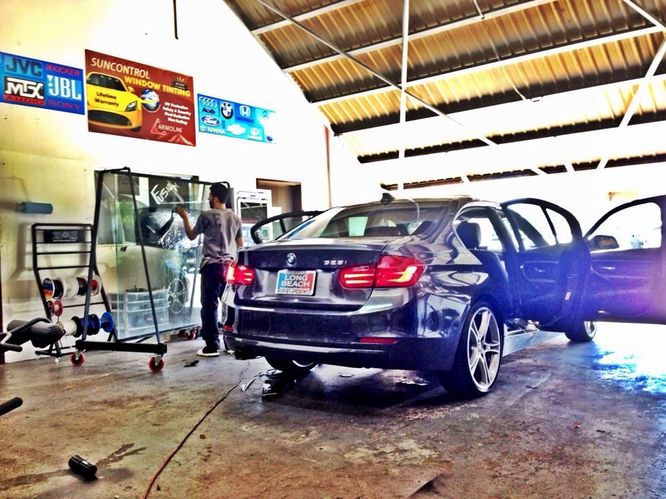 A dark sedan parked inside a garage workshop with its doors open as an individual works on a glass panel nearby.