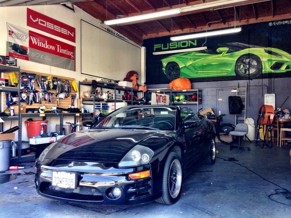 A black Mitsubishi Eclipse convertible parked inside an automotive workshop with wall banners and shelves of supplies.