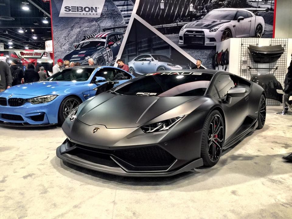 A matte black Lamborghini and a blue BMW displayed at a car show, with a large Seibon brand banner in the background.