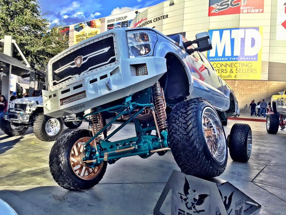 A lifted light blue pickup truck with large tires and a teal suspension sits on a display stand outside a convention.