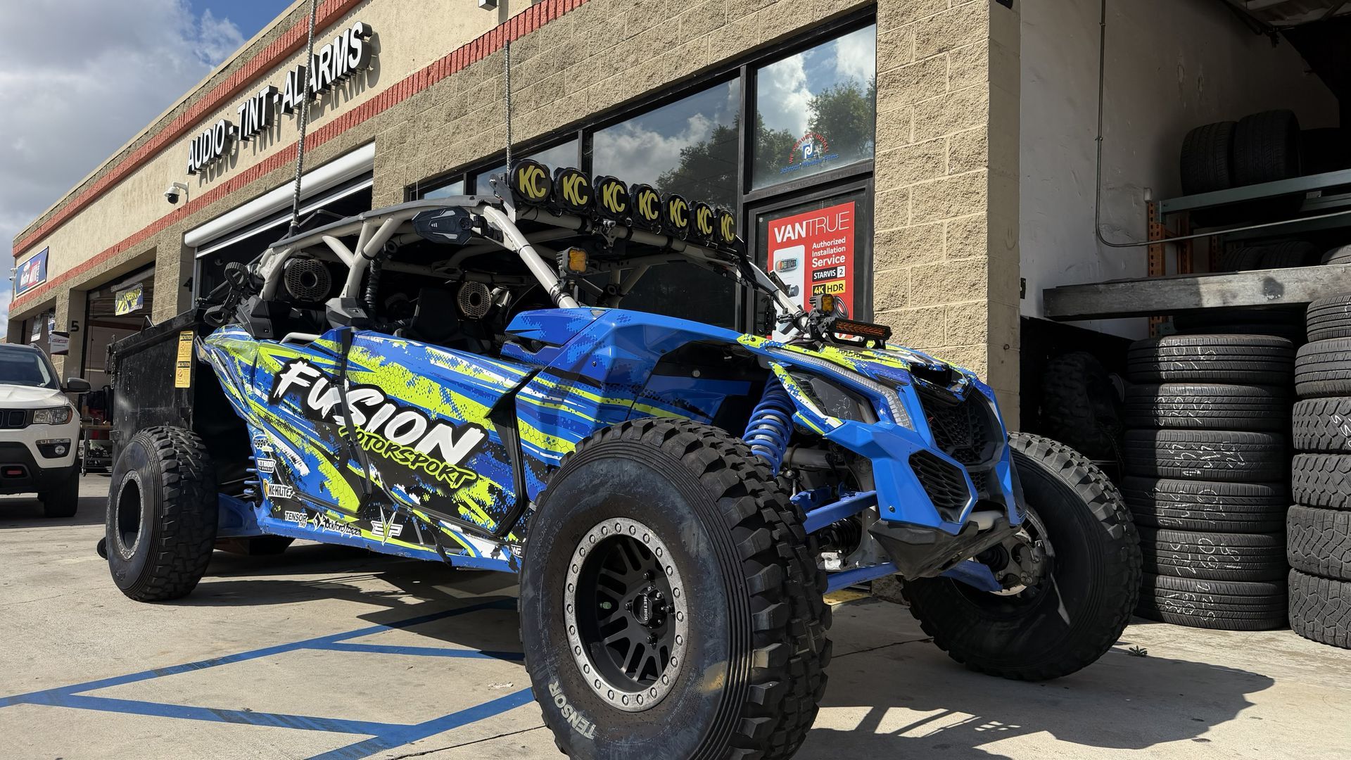 A blue, black, and green Fusion off-road racing vehicle parked outside a tire shop on a sunny day.