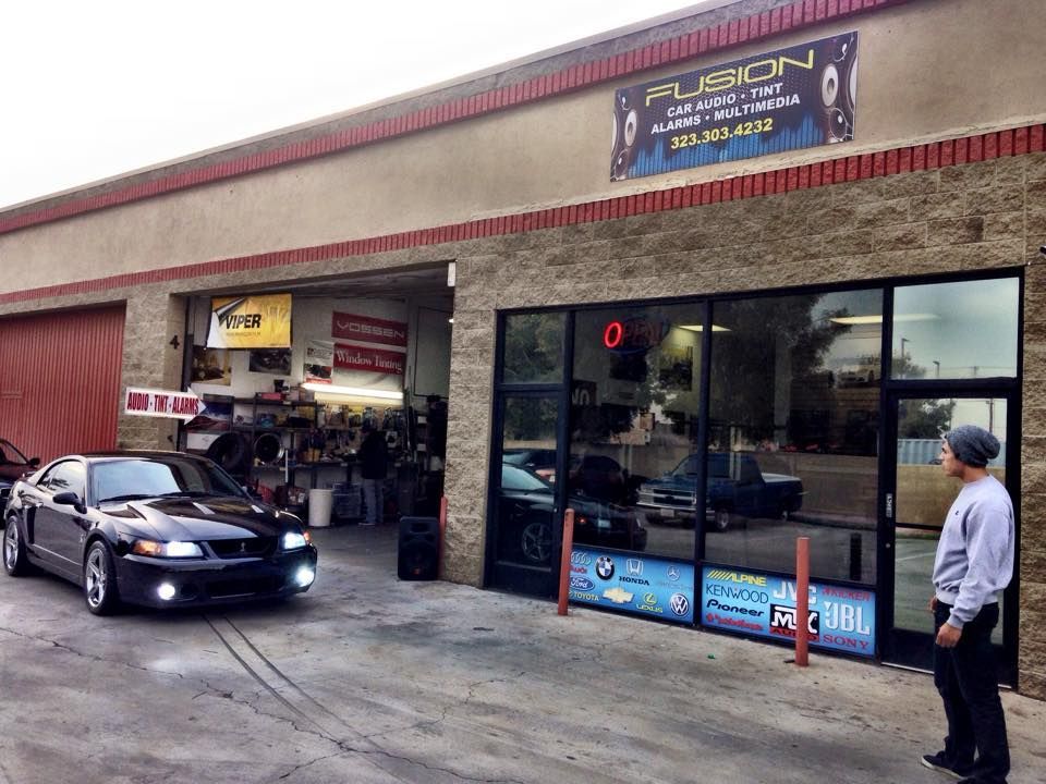 A black Ford Mustang sits outside an automotive shop called Fuson, with a person standing by the entrance.