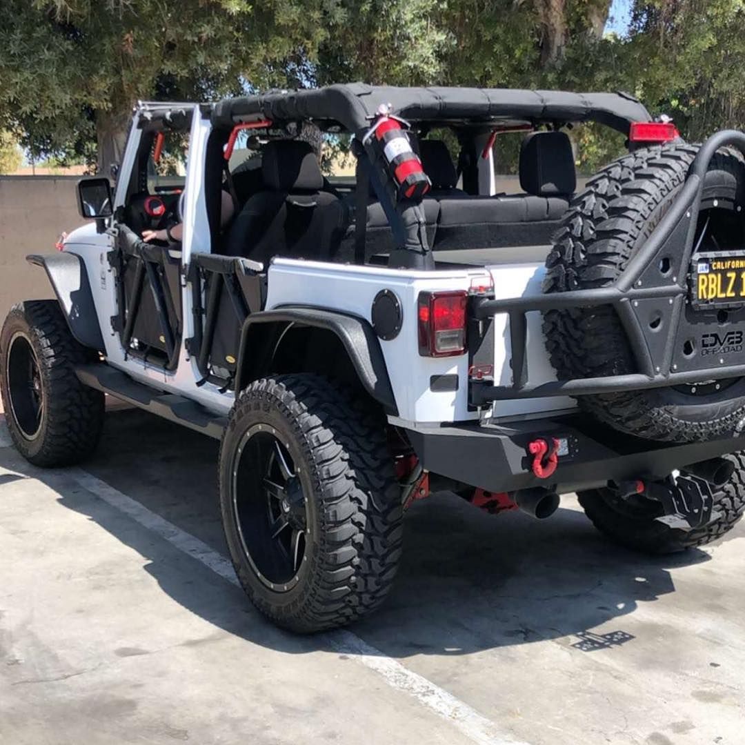 A white Jeep Wrangler with black off-road doors, large tires, and a rear-mounted spare tire parked on a concrete lot.