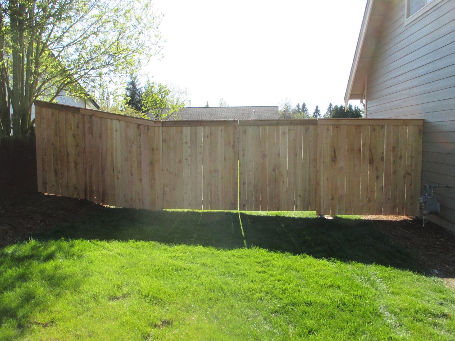 Wooden fence in a yard next to a house, with green grass in the foreground.