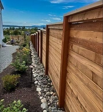Wooden fence with a rock bed along the side, plants and trees in the background, overlooking water.