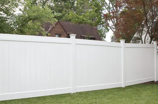 White vinyl privacy fence in front of a grassy yard, with a house and trees in the background.