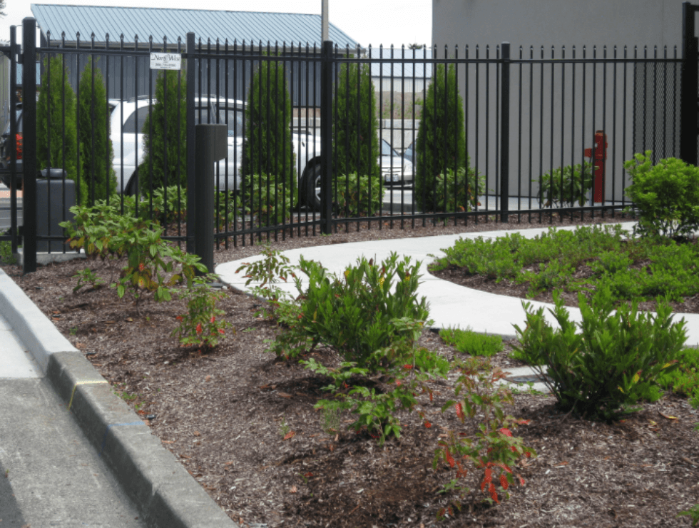 Black fence along a walkway with a flowerbed, bushes, and trees. White car visible behind the fence.
