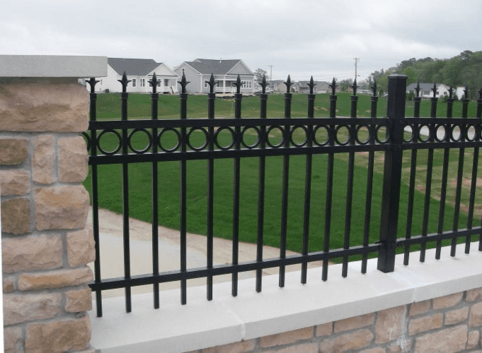 Black wrought-iron fence atop a stone wall, bordering a green lawn with houses in the background.