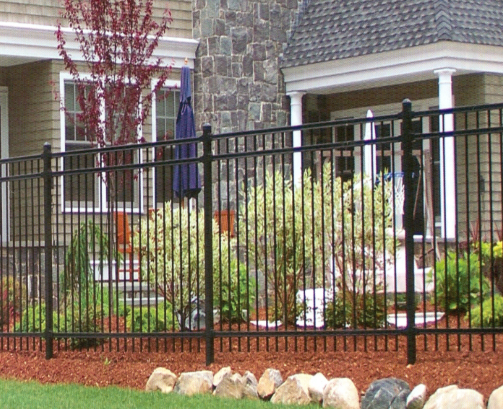 Black metal fence in front of a house with shrubs and red mulch.