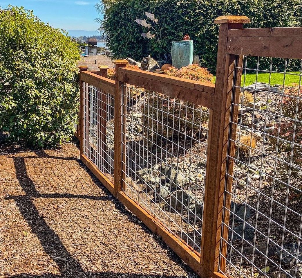 Wooden fence with wire mesh panels, separating a gravel area from a rock bed with green bushes and grass.
