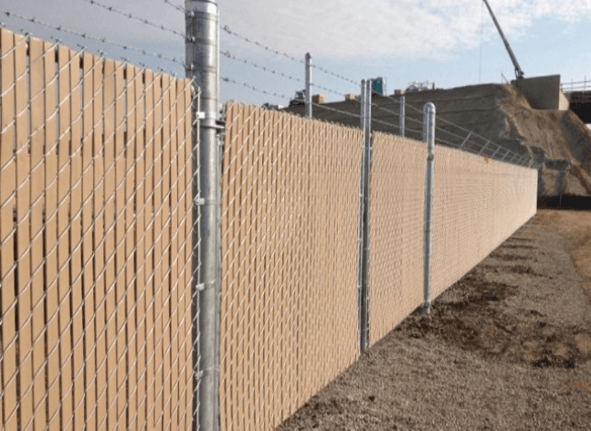 Tan privacy fence with barbed wire on top, along a construction site.