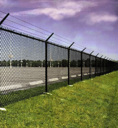 Black chain-link fence topped with barbed wire, bordering a paved area and grassy field under a cloudy sky.