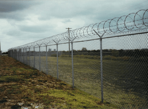 Chain-link fence with razor wire at the top, in a grassy field under a cloudy sky.