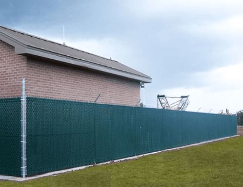 Green privacy fence around a brick building, topped with barbed wire under a cloudy sky.