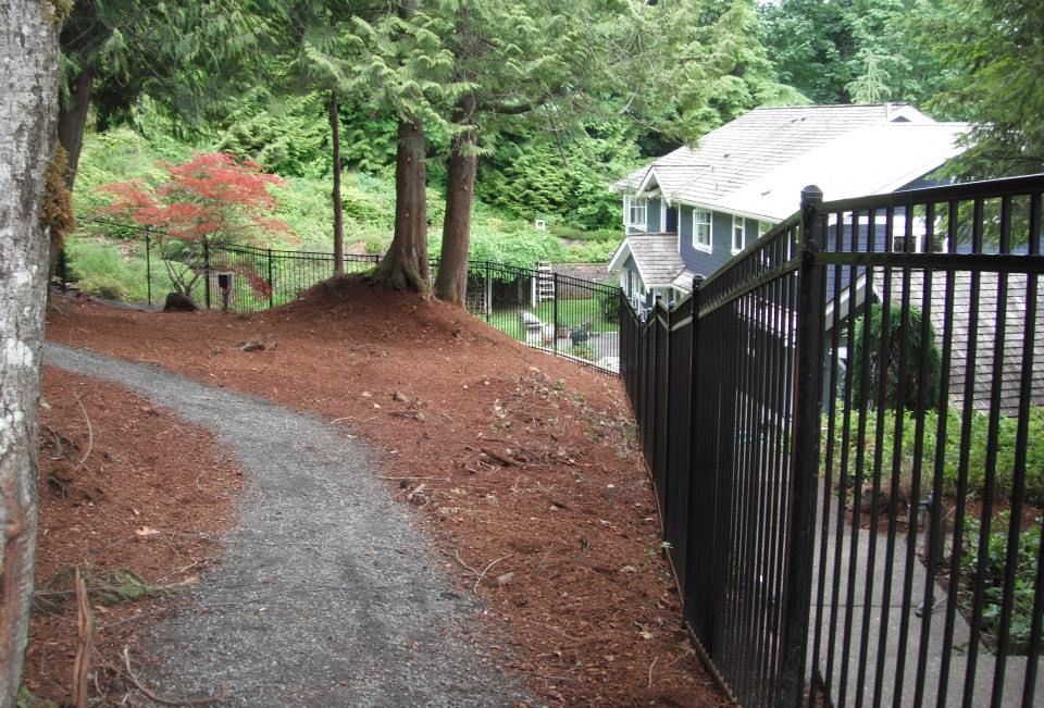A gravel path winds alongside a black fence, leading towards a house in a wooded area.