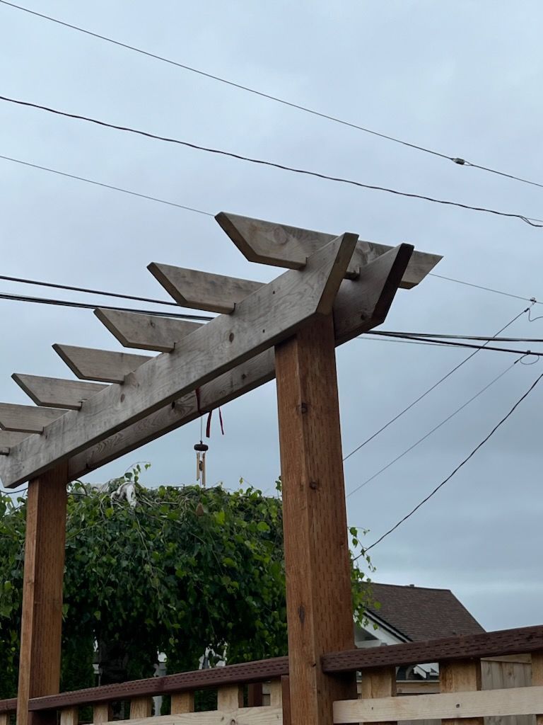 Wooden pergola with angled beams, brown support posts, against a cloudy sky and greenery.