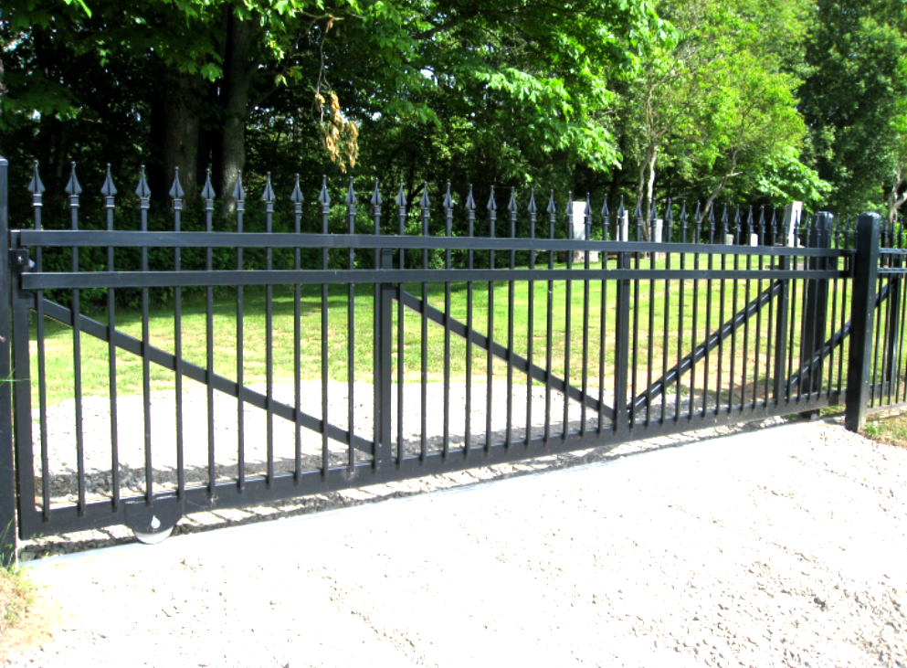Black metal gate with decorative top, spanning a gravel driveway, trees in background.