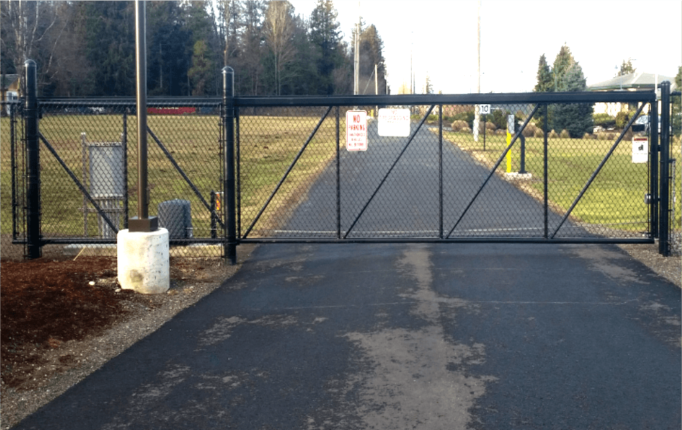 Black gate across a paved road, with a chain-link fence on either side, in front of a grassy area.