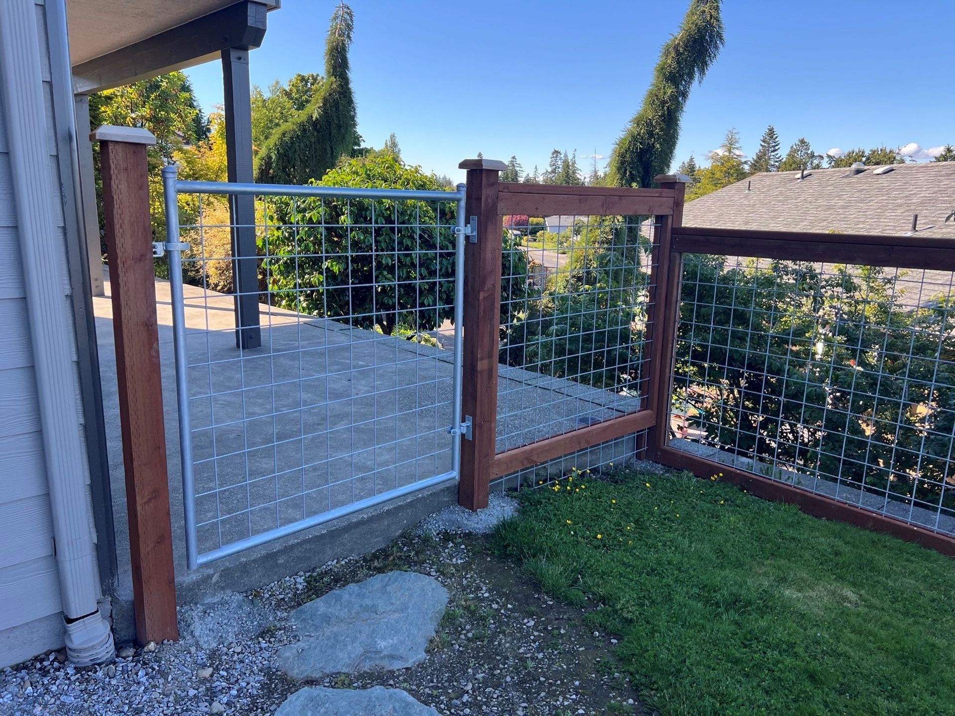 Gate with metal grid, wooden posts, gravel and grass, on a sunny day.