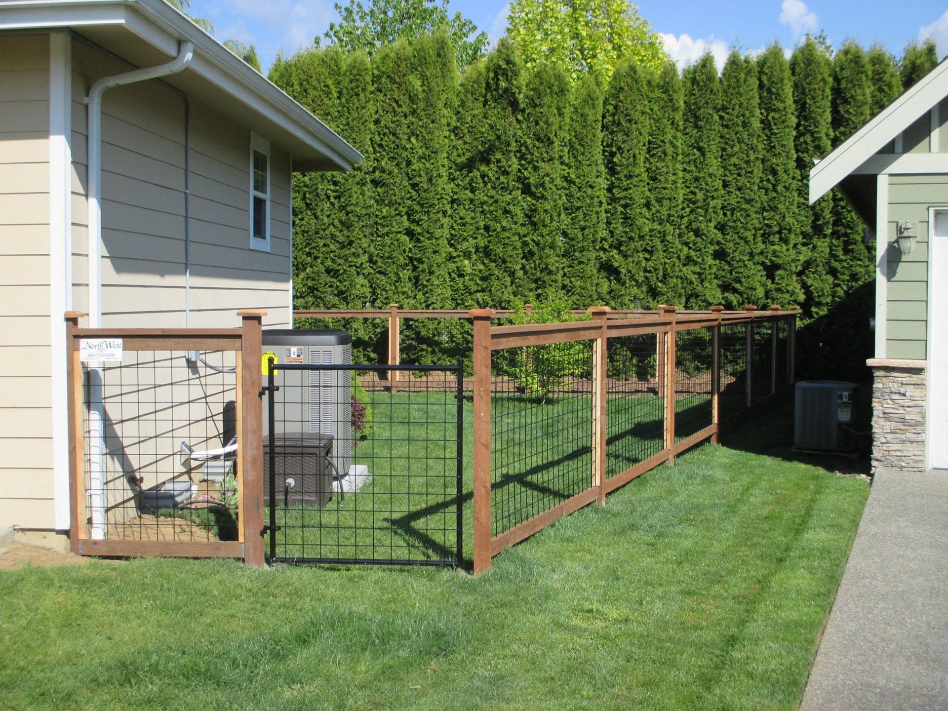 A wooden fence encloses an area near a house, separating it from the green lawn and evergreen trees in the background.