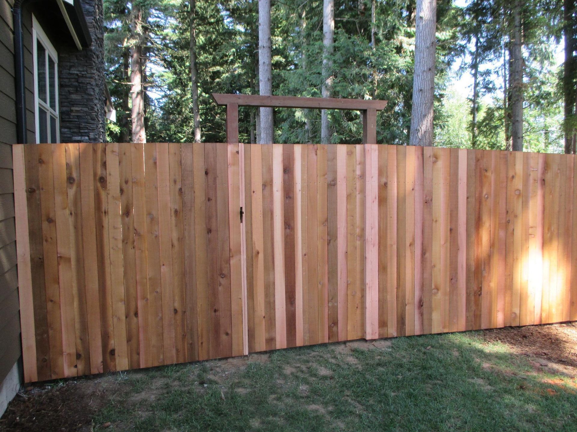 Wooden fence in a grassy backyard with a dark-colored house on the left and trees in the background.