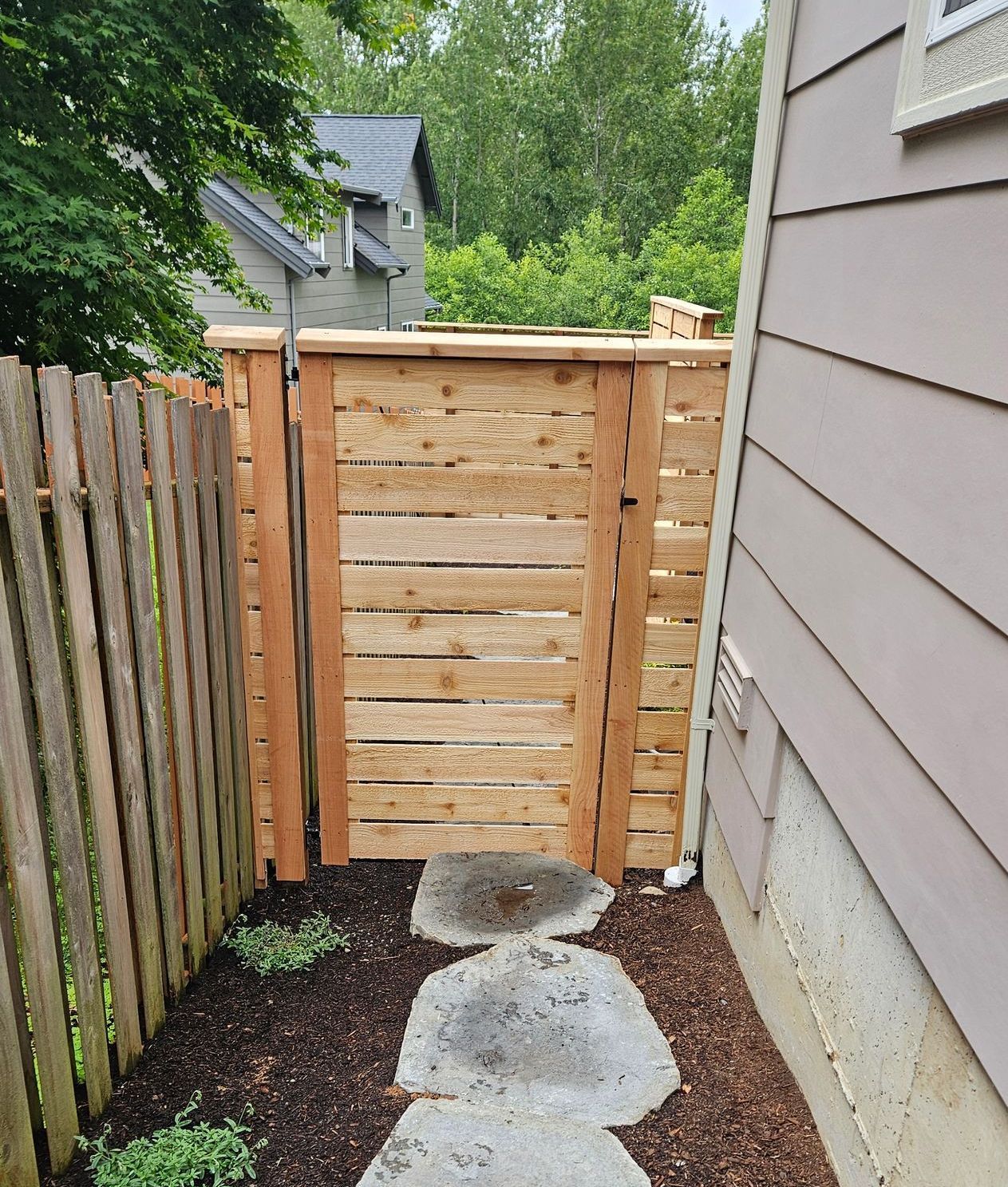 Wooden gate in a backyard with stepping stones leading toward it. The gate is between two fences and a house.