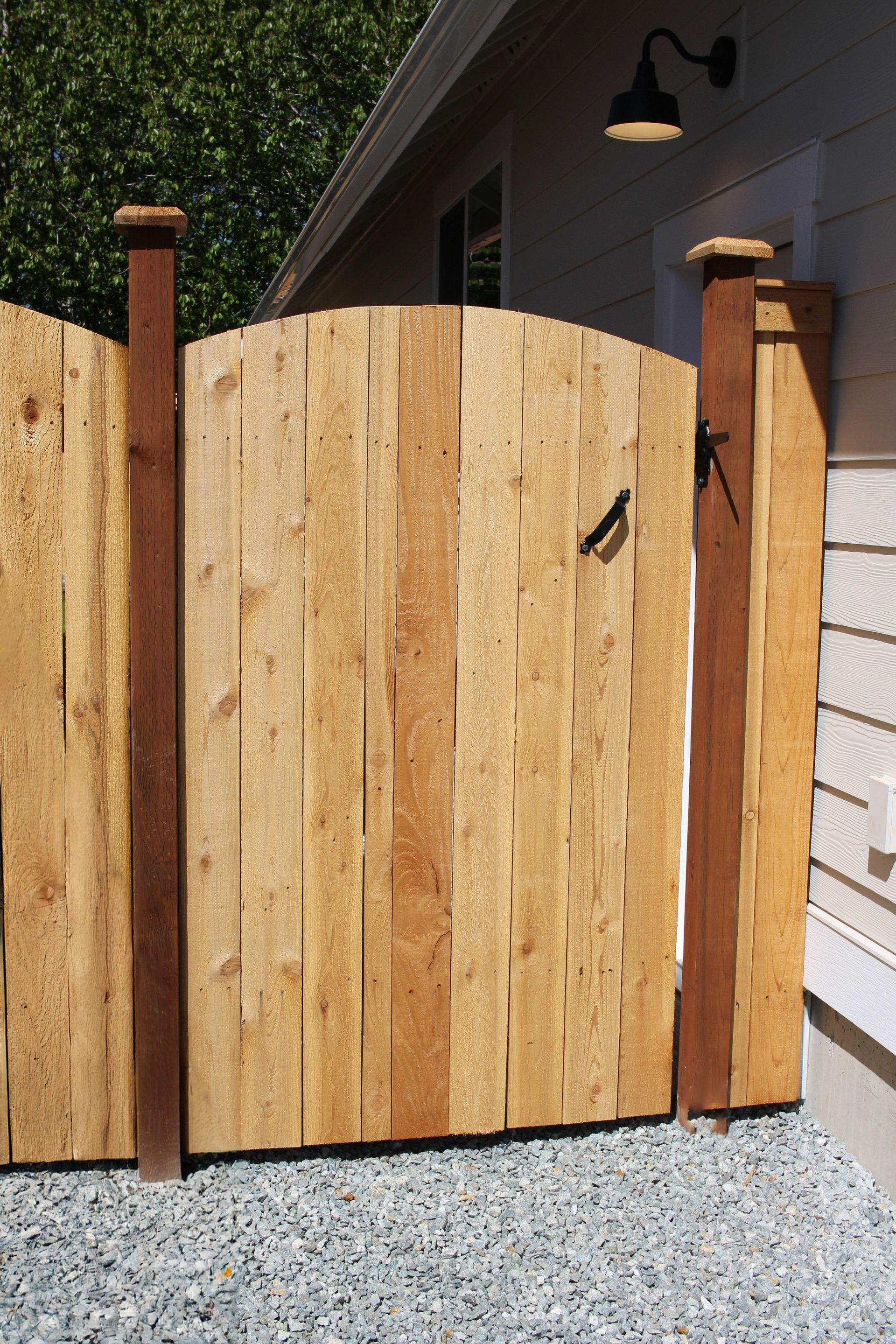 Wooden gate with arched top, between brown posts. Gate is on a gravel surface, next to a house.