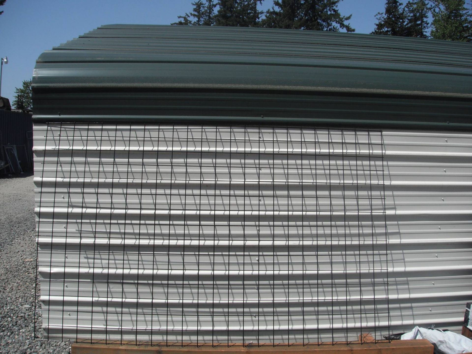 White corrugated metal building with dark vertical lines. Green roof, sunny outdoor setting.