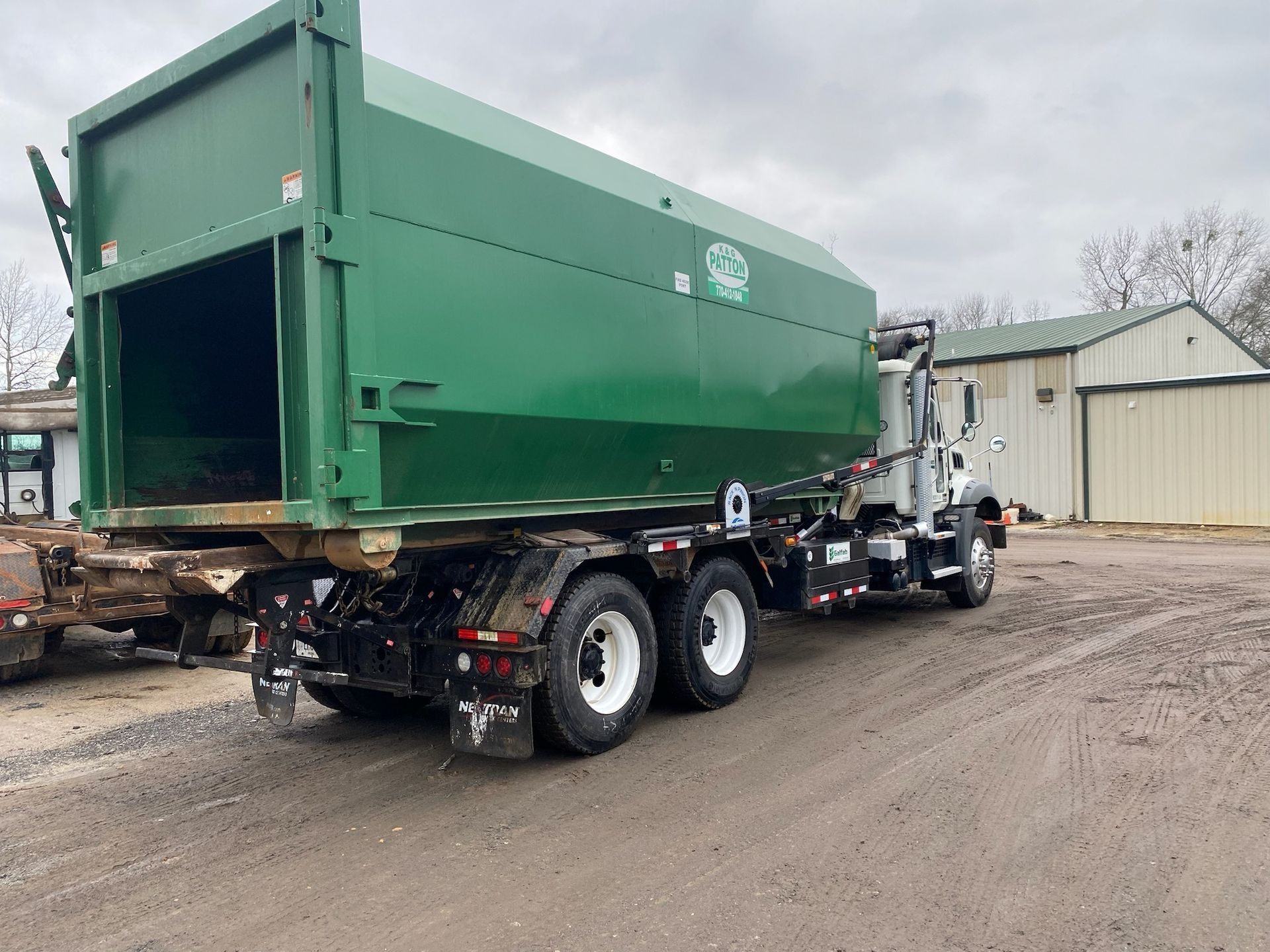 Green waste hauling truck parked on dirt lot near building.