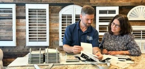 Two people review a sample book at a table in front of window shutters.
