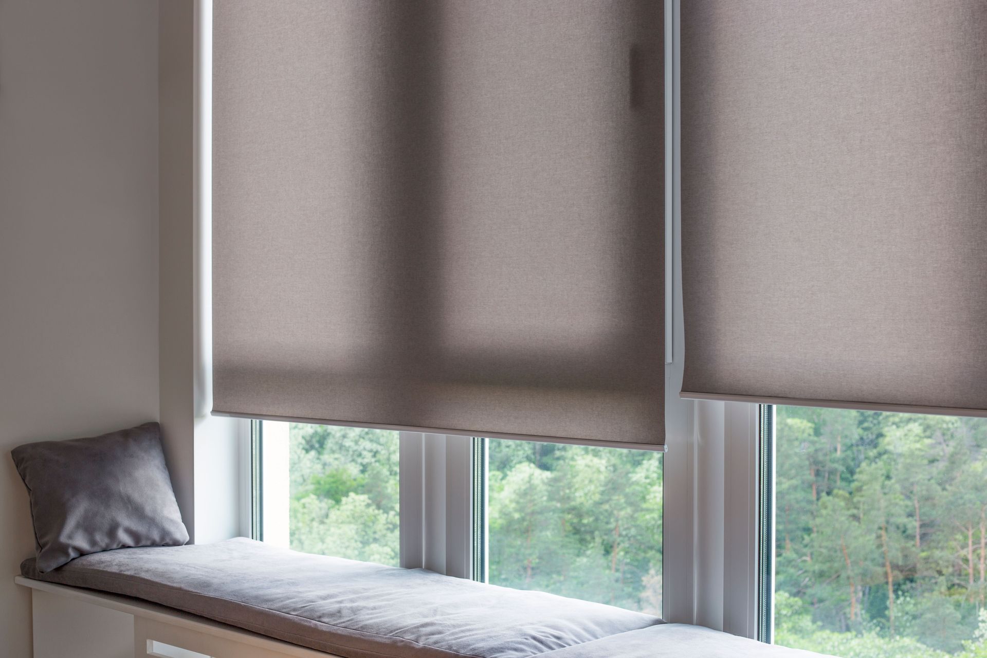 Window seat with beige blinds and a gray cushion, overlooking green trees outside