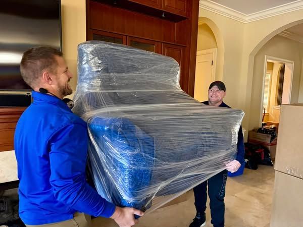 Two men are carrying a chair wrapped in plastic in a living room.