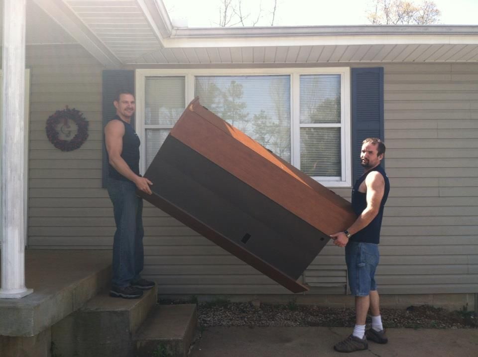 Two men are carrying a large piece of furniture in front of a house.