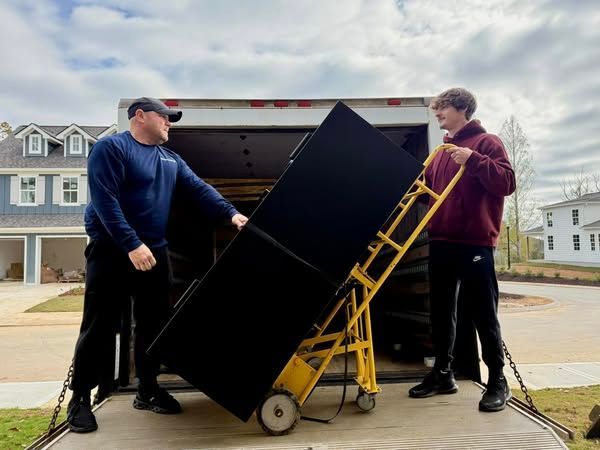 Two men are pushing a large black refrigerator on a dolly.