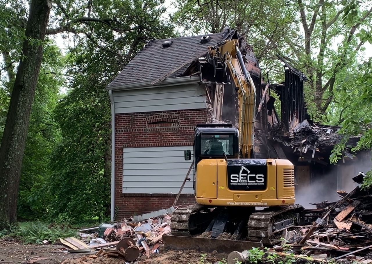 A yellow excavator is demolishing a brick house.