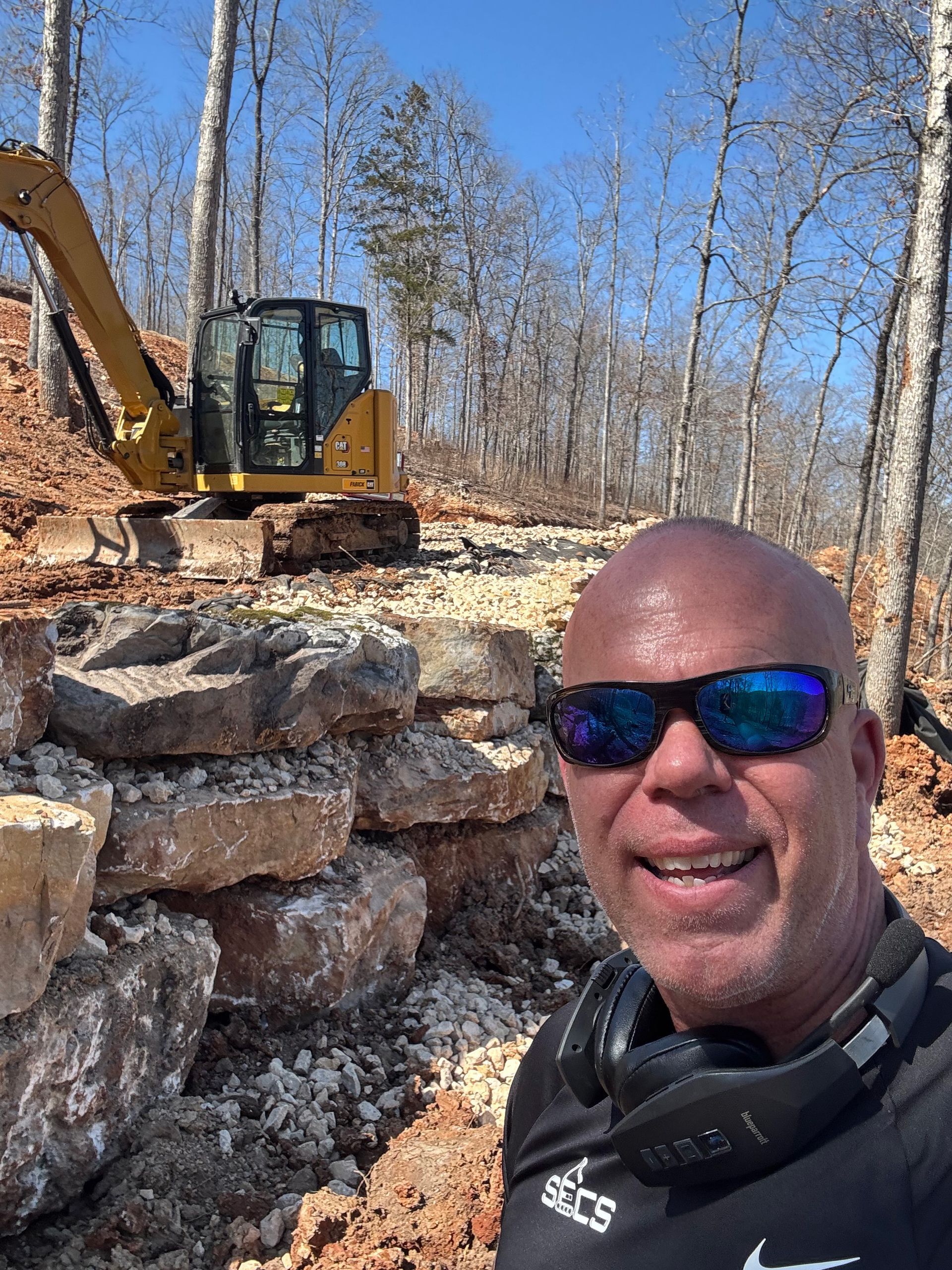 A man wearing sunglasses and headphones is standing in front of a bulldozer in the woods.