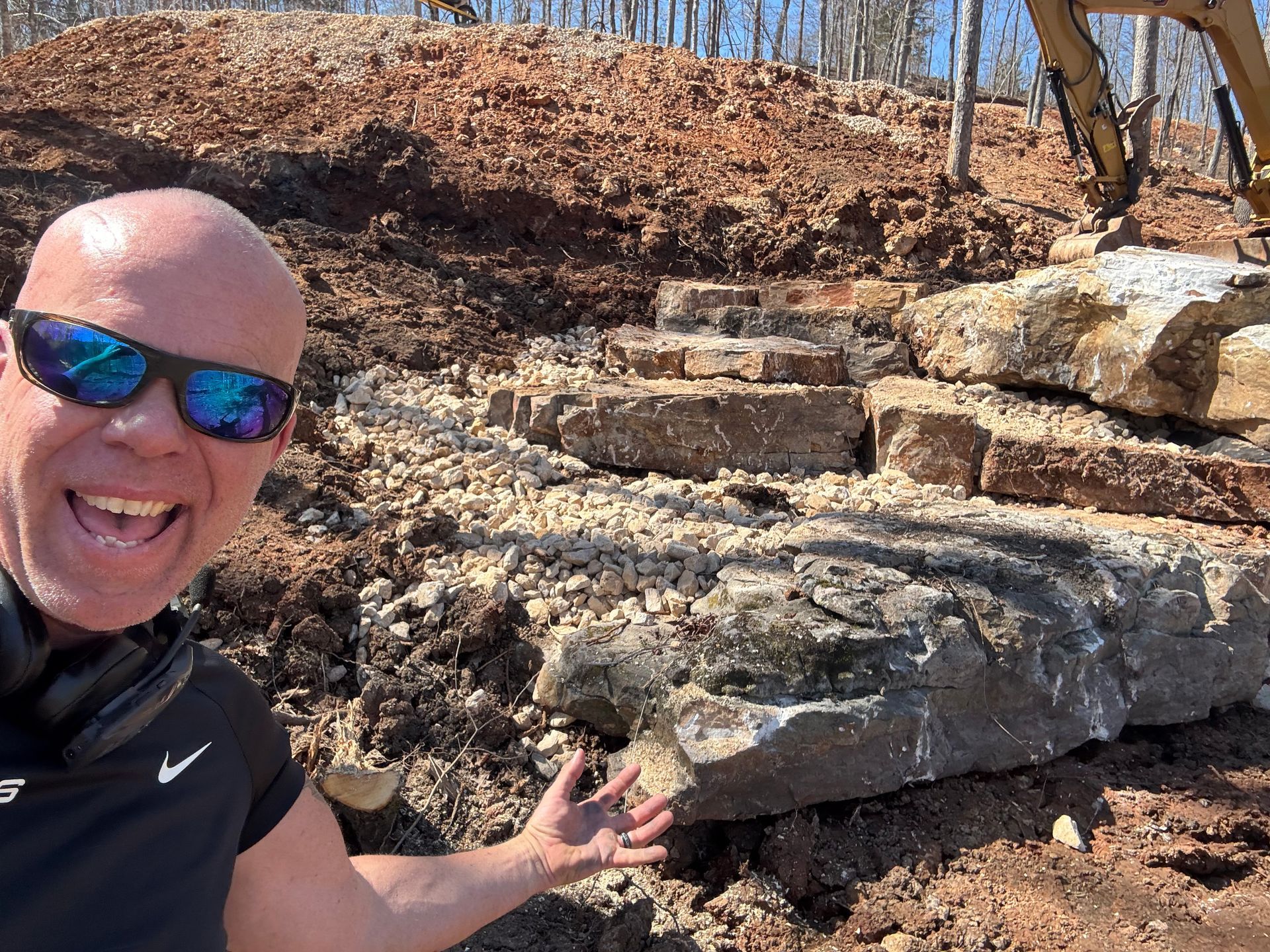 A bald man wearing sunglasses and headphones is standing in front of a pile of rocks.