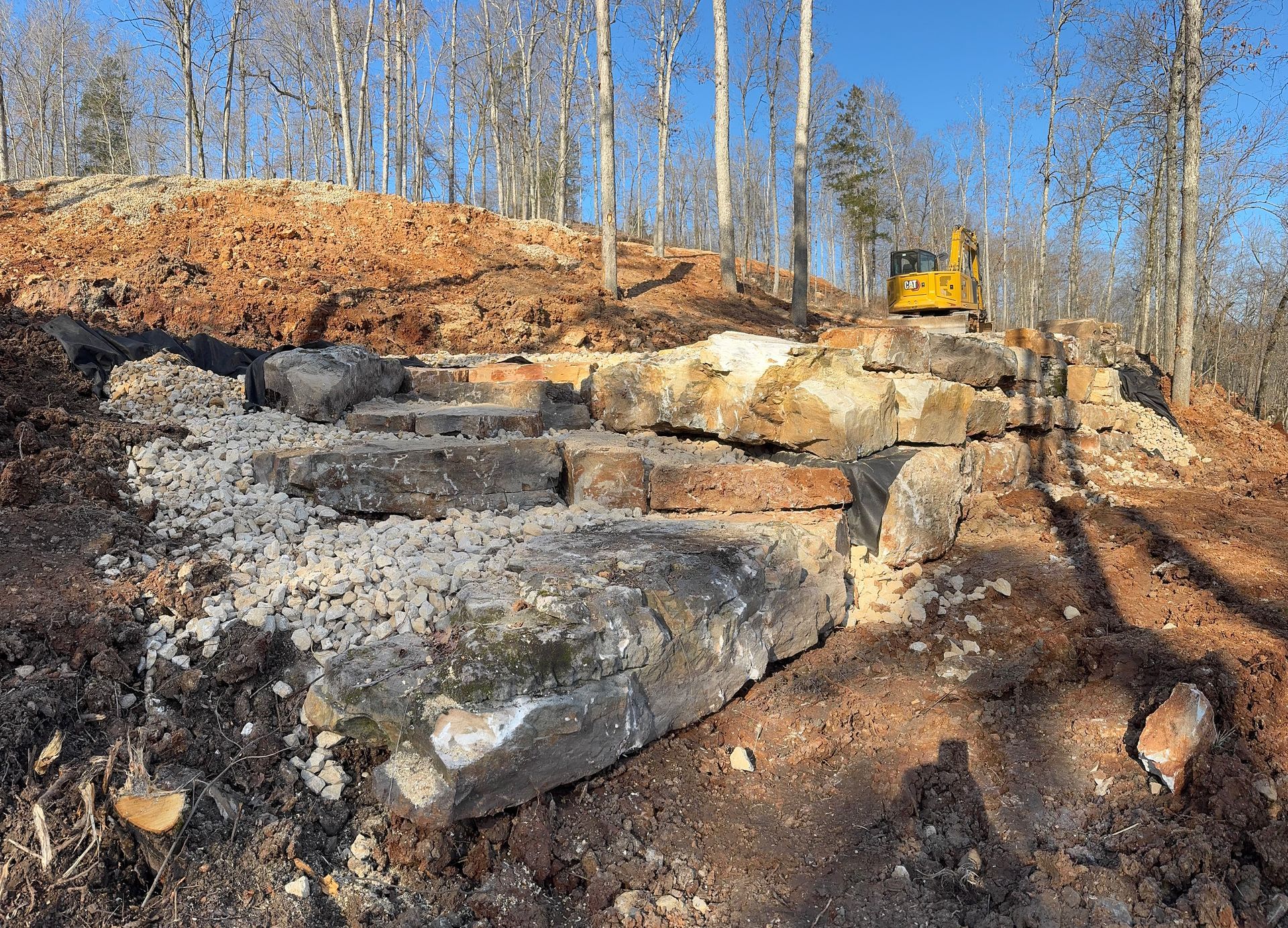 A yellow bulldozer is working on a rock wall in the woods.