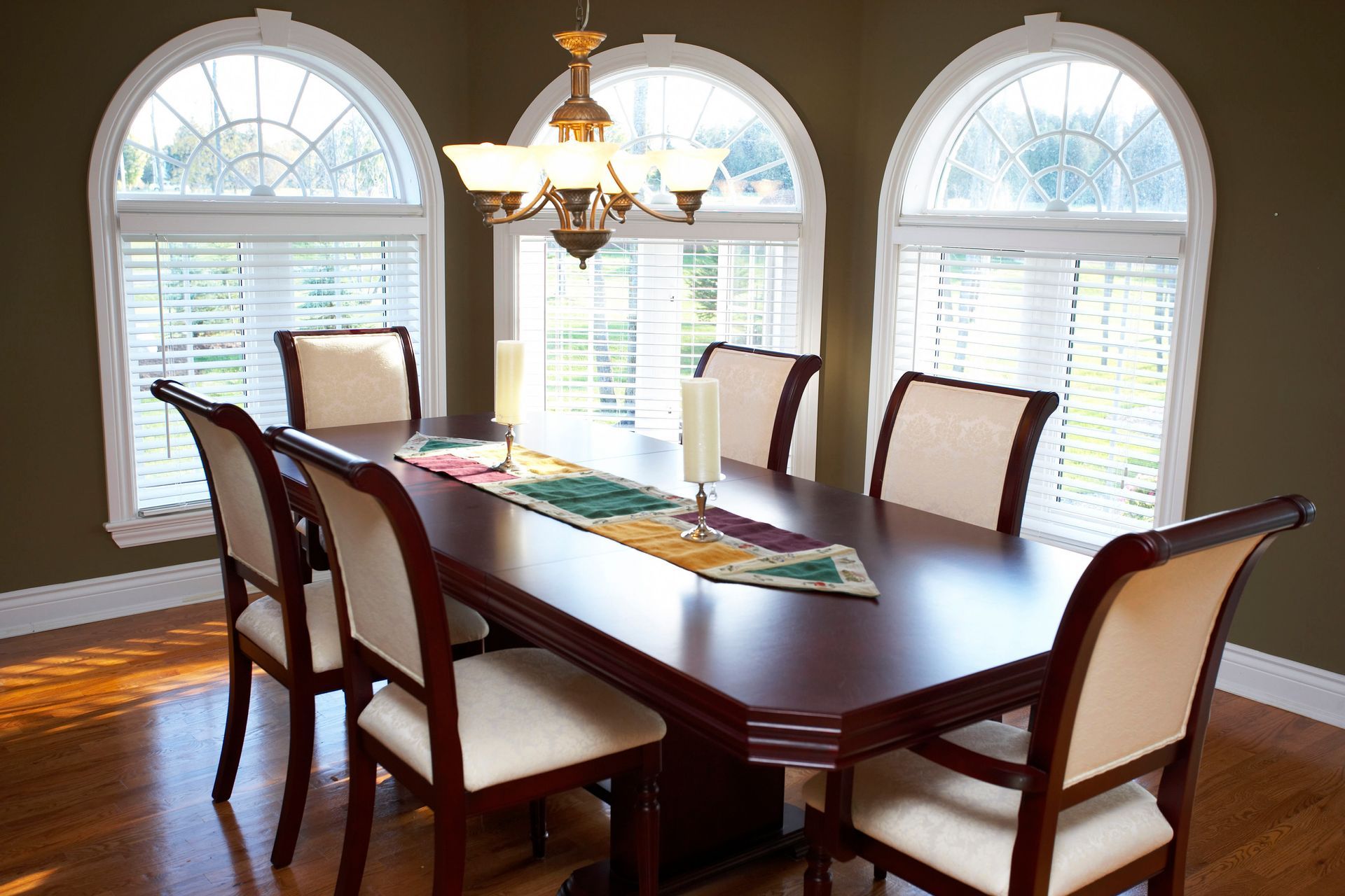 Dining room with a dark wood table, upholstered chairs, chandelier, and two arched windows with blinds.