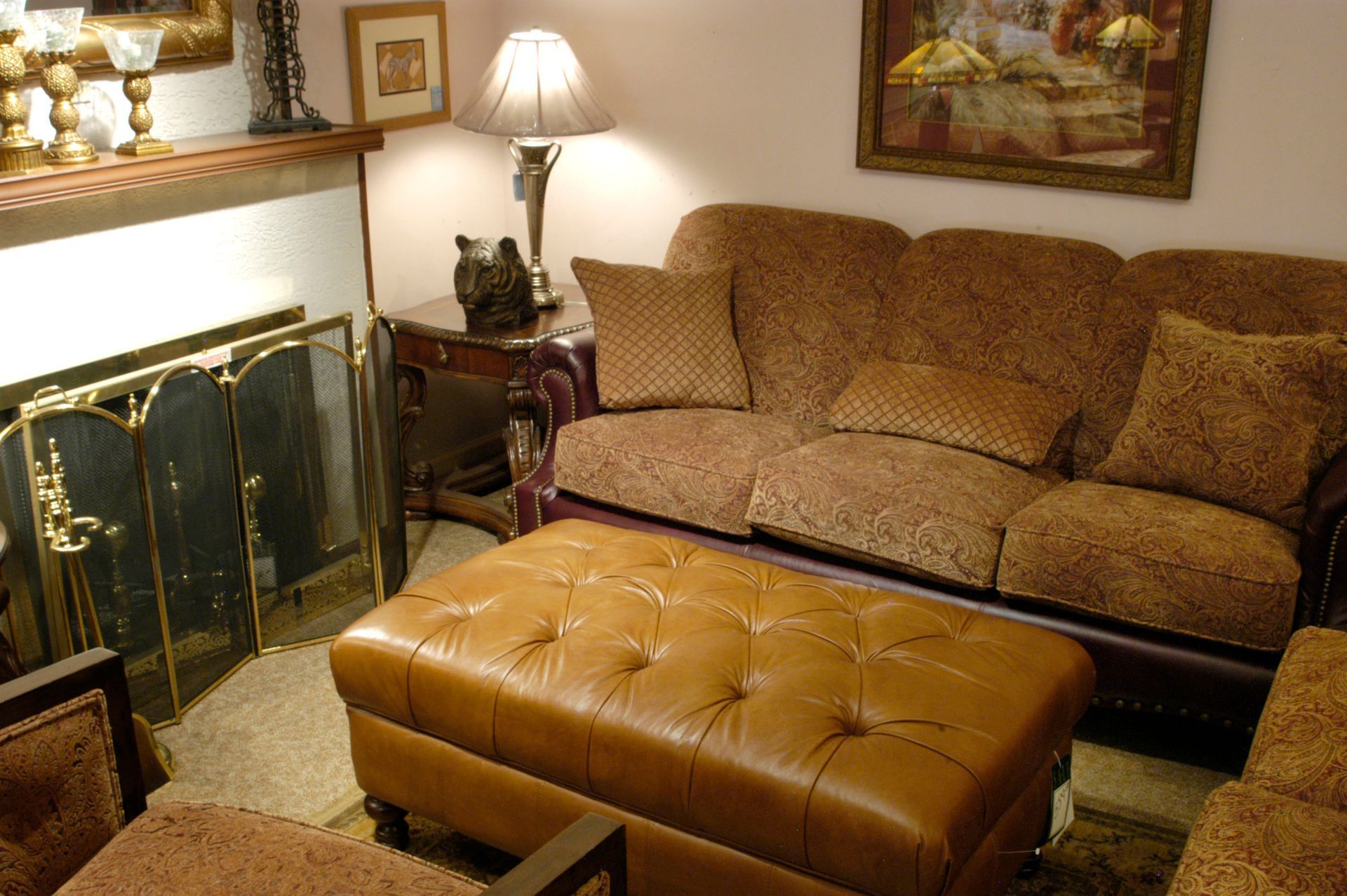 Cozy living room with a brown sofa, tufted ottoman, fireplace, lamp, and framed wall art.