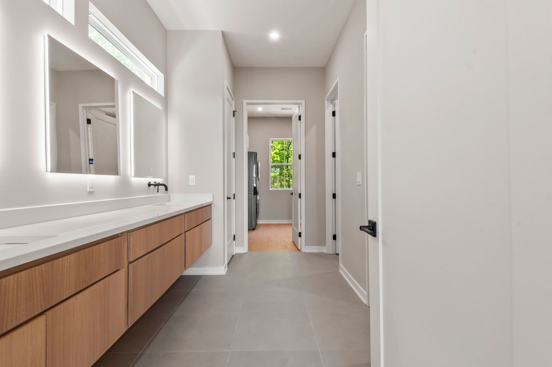 Modern bathroom vanity with wood cabinets, large mirror, and a hallway leading to a bright doorway