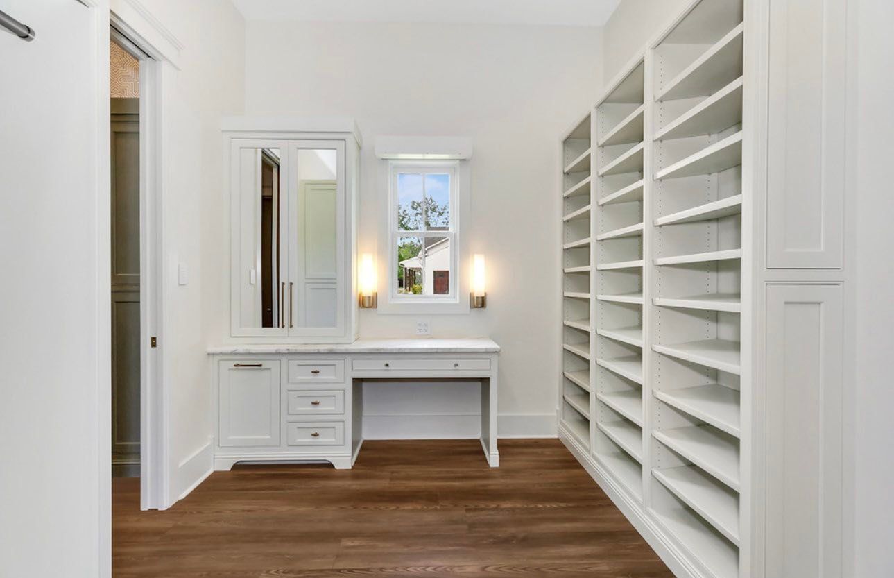 Bright white walk-in closet with built-in shelves, a desk, mirror, and wood floor.