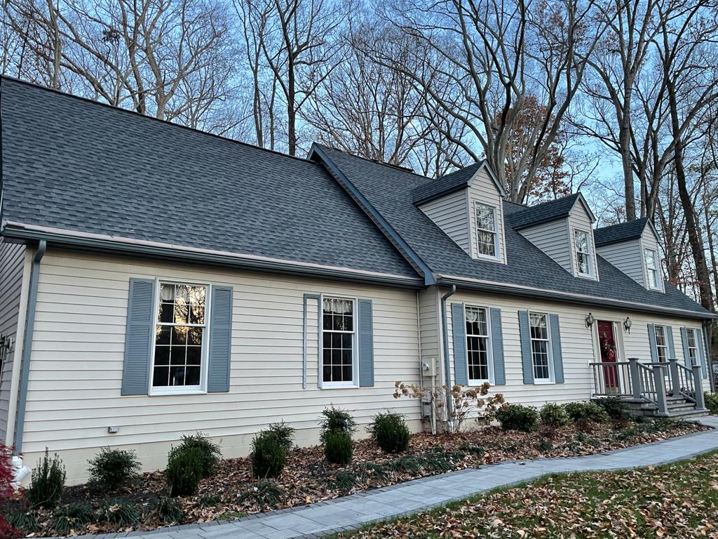 A large white house with a gray roof and shutters is surrounded by trees.